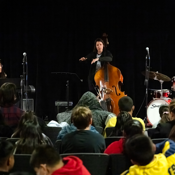Bass player presenting at Jazz for the Schools, photo is taken facing stage over a sea of students.
