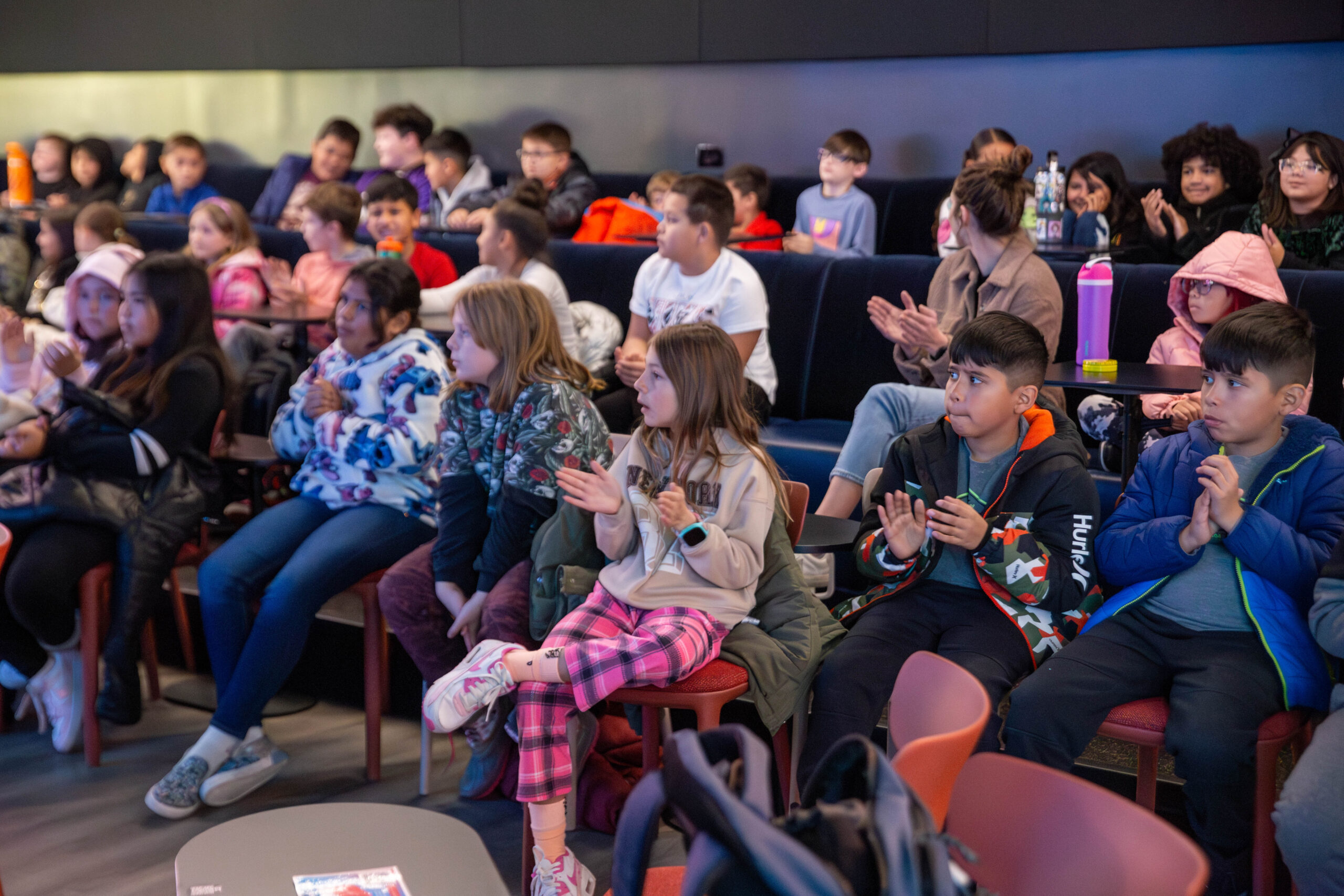 Elementary school students clap along during a Jazz for the Schools presentation, crafted by Gift of Jazz and hosted at Dazzle Denver. Many rows of students clap and watch the stage. 